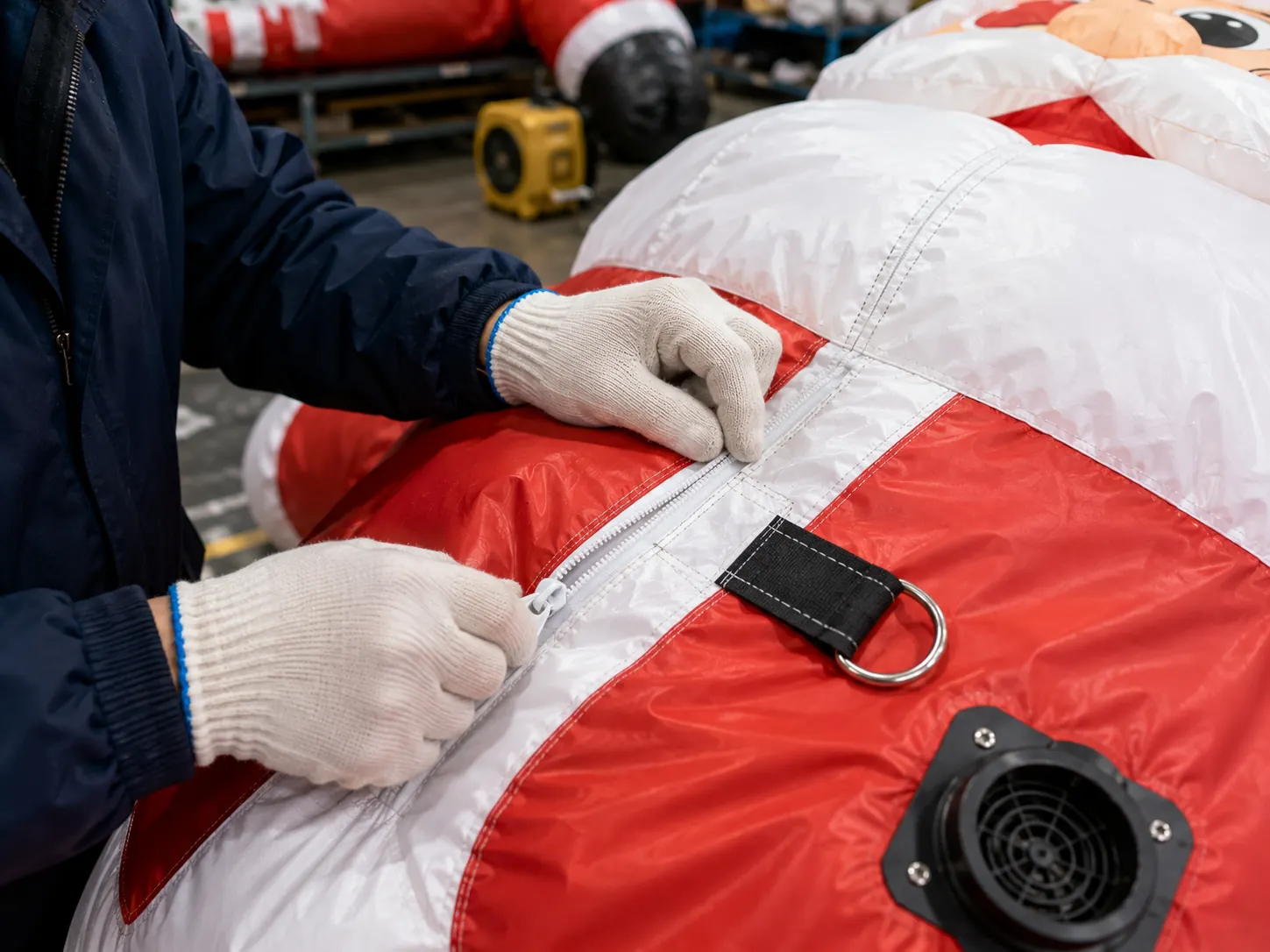 Gloved technician inspecting a reinforced zipper seam, anchor ring and blower inlet on a red and white inflatable Christmas display
